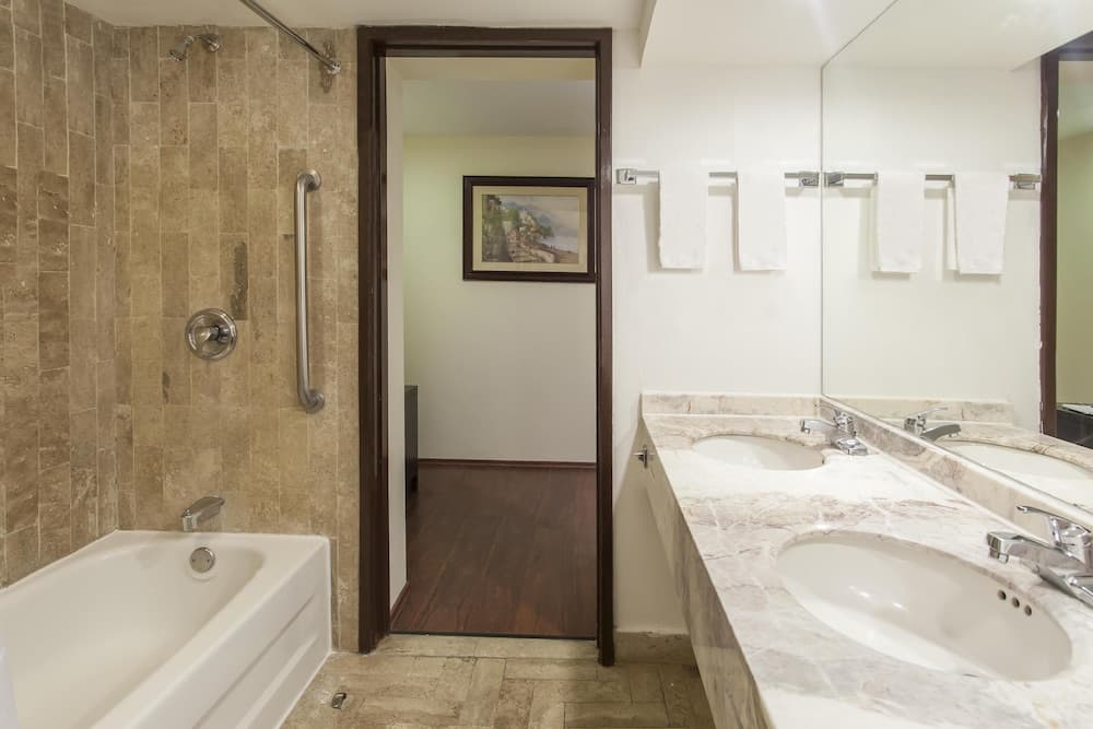 A cozy hotel bathroom features a tiled bathtub, marble countertop with dual sinks, neatly arranged towels, and a serene view into an adjacent room with warm wood flooring and framed artwork, ideal for travelers seeking comfort and cleanliness.