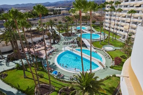 A vibrant resort scene featuring lush palm trees, sparkling blue pools, and modern buildings under sunny skies, ideal for a relaxing vacation getaway.