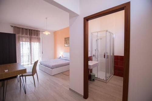 A cozy studio apartment features a neatly made bed by a window, a small dining area, and an open bathroom with a glass shower, ideal for travelers seeking comfort in a compact space.
