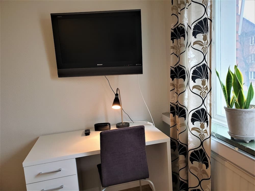 A cozy home office setup features a white desk with a purple chair, a mounted TV, a desk lamp, and a potted plant by the window, ideal for travelers seeking a comfortable workspace during their stay.