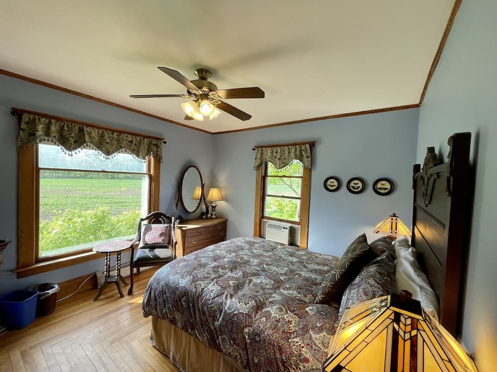 A cozy bedroom with wooden floors, two windows overlooking green fields, a patterned bedspread, and vintage decor creates a serene retreat ideal for travelers seeking a peaceful countryside stay.