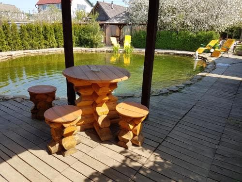 A serene garden deck features rustic log furniture beside a tranquil pond, surrounded by blooming trees and vibrant greenery, ideal for a peaceful retreat or family vacation spot.