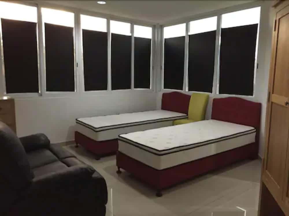 A cozy guest room features two single beds with colorful cushions, a leather armchair, and large windows with black blinds, ideal for travelers seeking comfort and privacy.