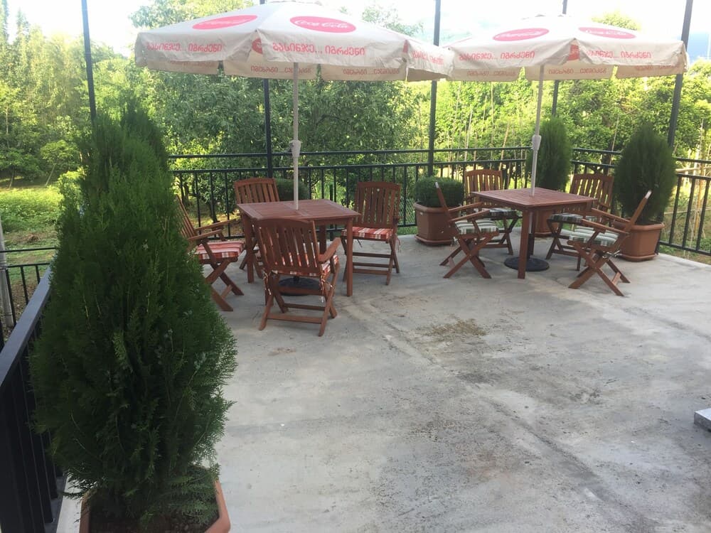 A serene outdoor patio features wooden tables and chairs under Coca-Cola branded umbrellas, surrounded by lush greenery, ideal for a relaxing dining experience in nature.