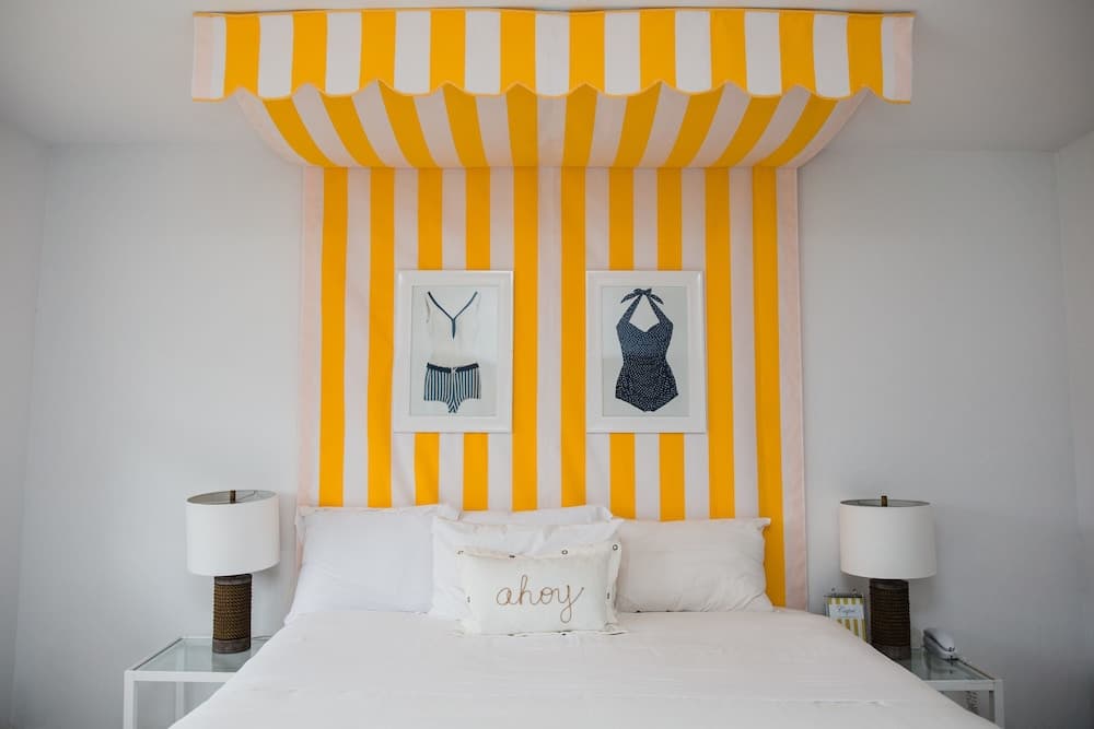 A vibrant bedroom features a striking yellow-and-white striped canopy over a cozy bed adorned with "ahoy" pillows, framed swimwear art, and bedside lamps, ideal for travelers seeking a cheerful and stylish retreat.
