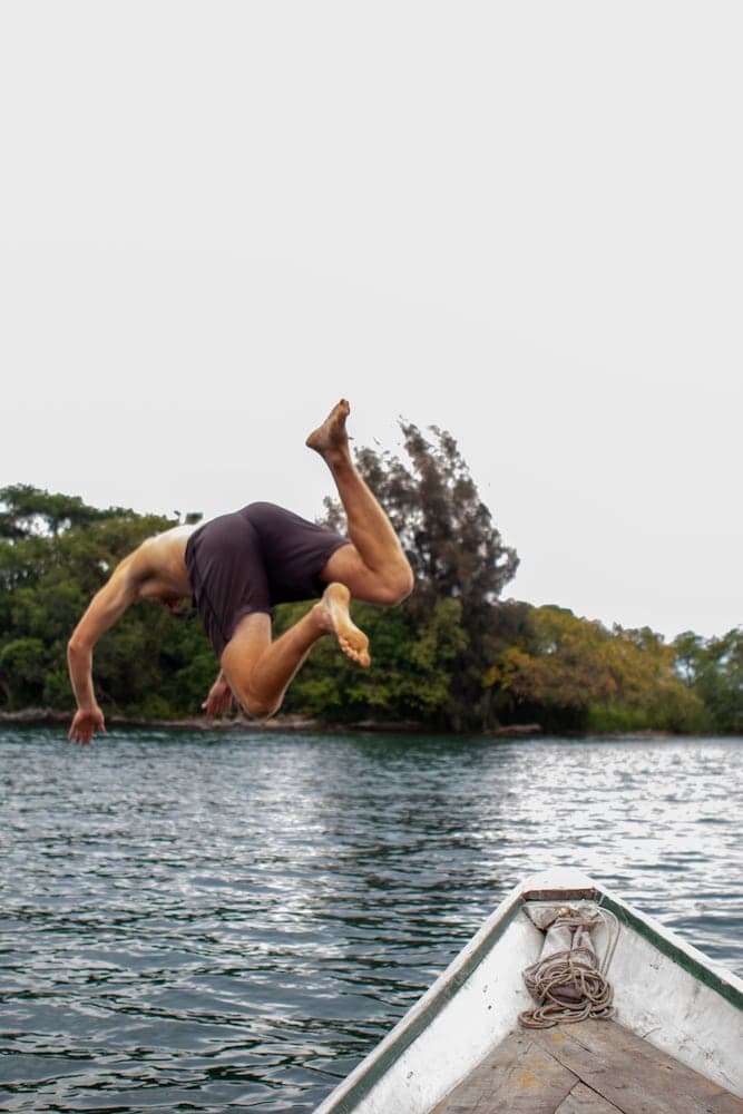 A daring traveler performs an acrobatic flip off a wooden boat into serene waters surrounded by lush greenery, capturing the thrill of adventure in a tranquil lakeside setting.