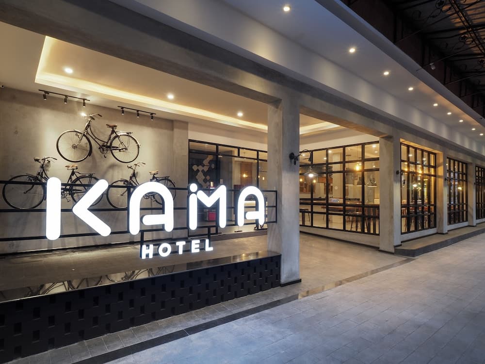 A modern hotel entrance featuring a glowing "KAMA HOTEL" sign, bicycles as decor, and large glass windows, creating an inviting urban atmosphere ideal for travelers seeking stylish accommodations.