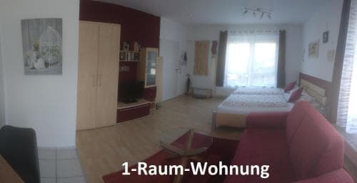 A cozy studio apartment features a bright living space with a red sofa, wooden furniture, and large windows, ideal for short-term travelers seeking comfort in a compact urban setting.