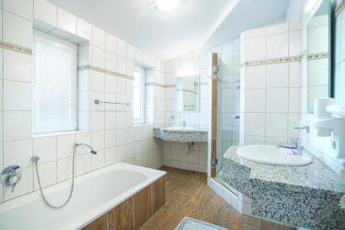 A bright, modern bathroom with white tiled walls, wooden flooring, a bathtub, double sinks, and ample natural light through windows, ideal for travelers seeking comfort and cleanliness in their accommodations.