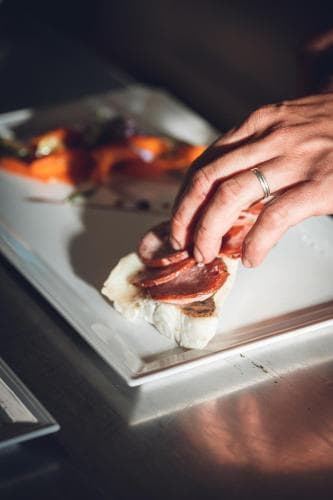 A chef delicately places slices of cured meat onto a creamy appetizer on a sleek white plate, capturing the intimate ambiance of a gourmet dining experience ideal for culinary enthusiasts.