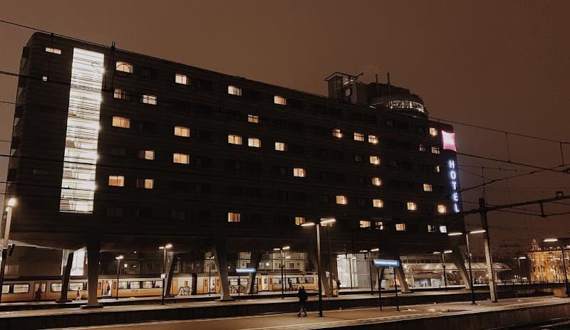 A warmly lit hotel building stands above a bustling train station at night, creating an inviting urban atmosphere ideal for travelers seeking convenient accommodation near Amsterdam's central transportation hub.