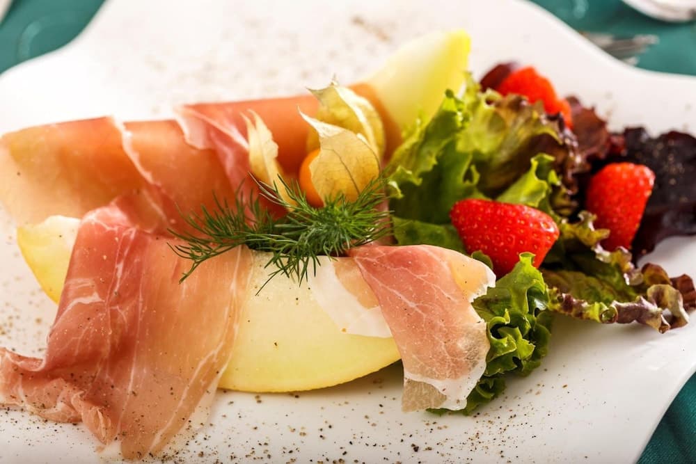 A beautifully plated dish featuring prosciutto-wrapped melon slices garnished with fresh herbs, accompanied by a vibrant salad of strawberries and lettuce, evokes a refined dining experience perfect for culinary travelers seeking gourmet delights.
