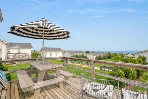 A serene coastal deck features a striped umbrella over a wooden picnic table, surrounded by lush greenery and distant houses, ideal for relaxing beachside getaways.