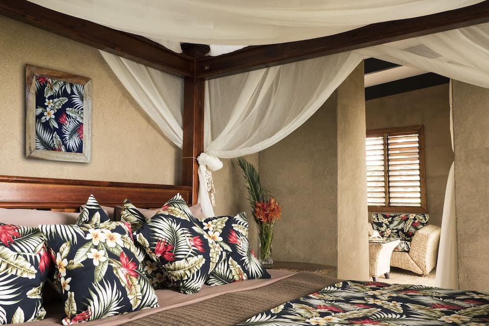 A cozy tropical-themed bedroom with a four-poster bed adorned in floral patterns, soft lighting through wooden shutters, and a relaxed ambiance perfect for a serene vacation retreat.
