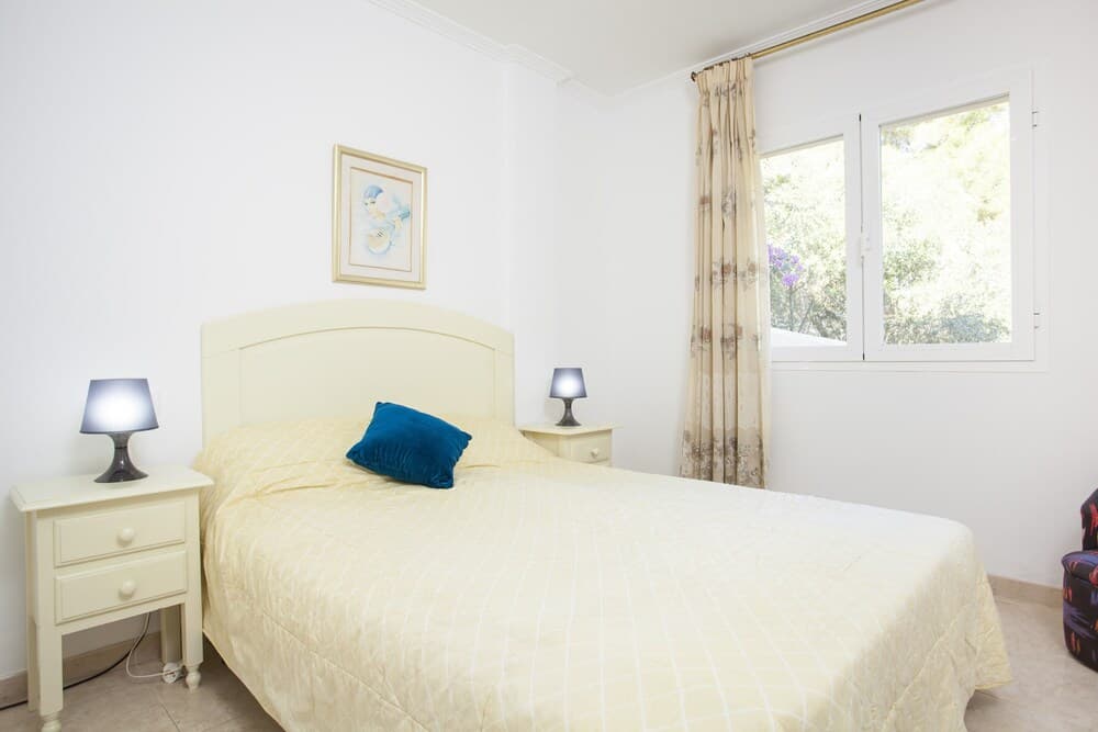 A serene bedroom with a cozy bed adorned in yellow linens, framed artwork, and natural light streaming through a window with floral curtains, ideal for travelers seeking a peaceful retreat.