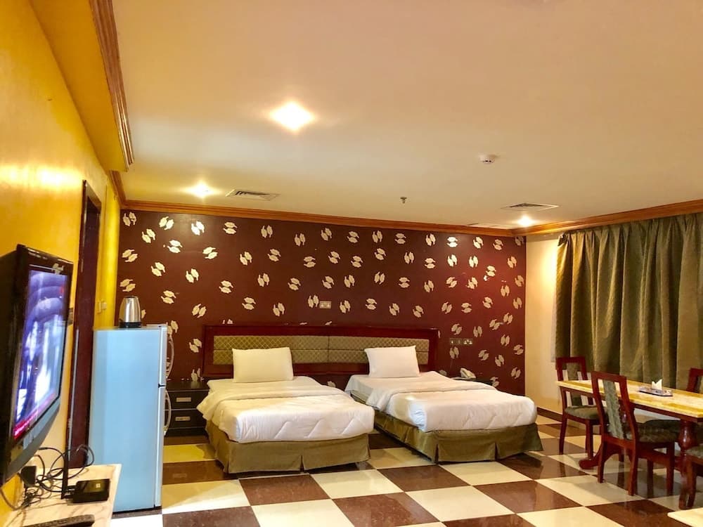 A cozy hotel room features twin beds against a patterned wall, checkered flooring, a dining area, and amenities like a TV and mini-fridge, ideal for travelers seeking comfort and convenience.
