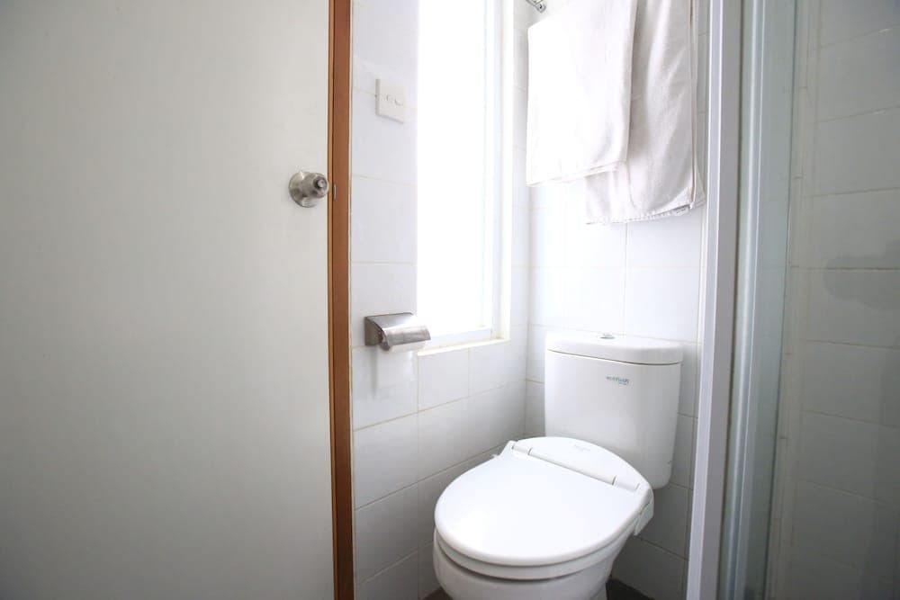 A clean, minimalist bathroom with white tiles, a toilet, a towel hanging neatly, and natural light from a window, ideal for travelers seeking simple accommodations.