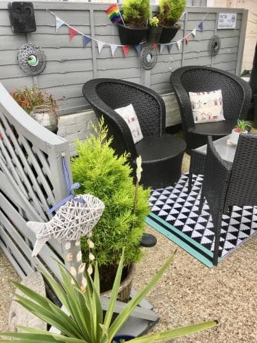 A cozy backyard patio adorned with festive bunting, black wicker chairs, vibrant greenery, and decorative elements creates an inviting outdoor retreat perfect for relaxation or small gatherings.