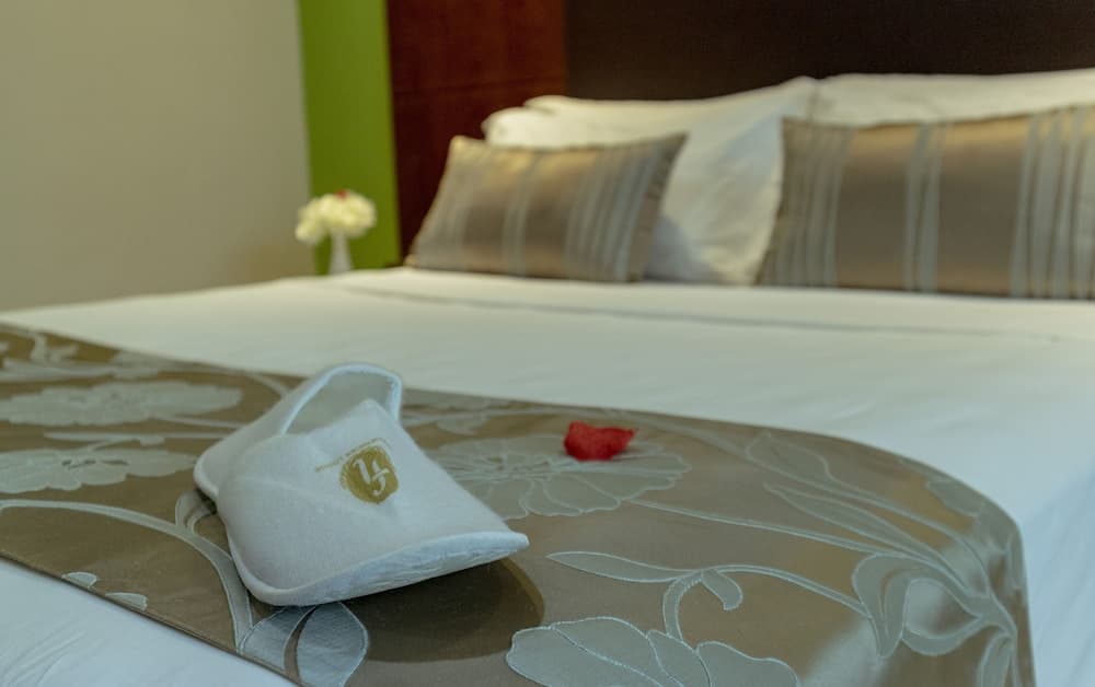 A cozy hotel room exudes comfort with its neatly made bed adorned with plush pillows, a decorative throw blanket featuring a white slipper and red rose petal, and a vase of flowers adding a touch of elegance, perfect for travelers seeking relaxation.