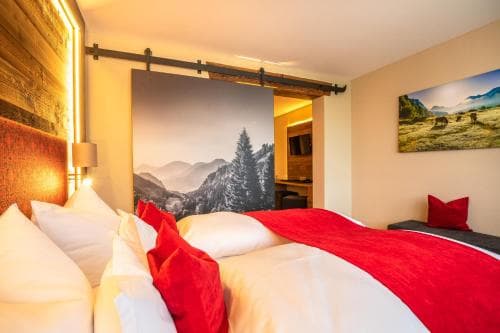 A cozy hotel room features a neatly made bed with red accents, a rustic wooden wall, and mountain-themed decor, creating a warm and inviting atmosphere ideal for travelers seeking relaxation in a scenic location.