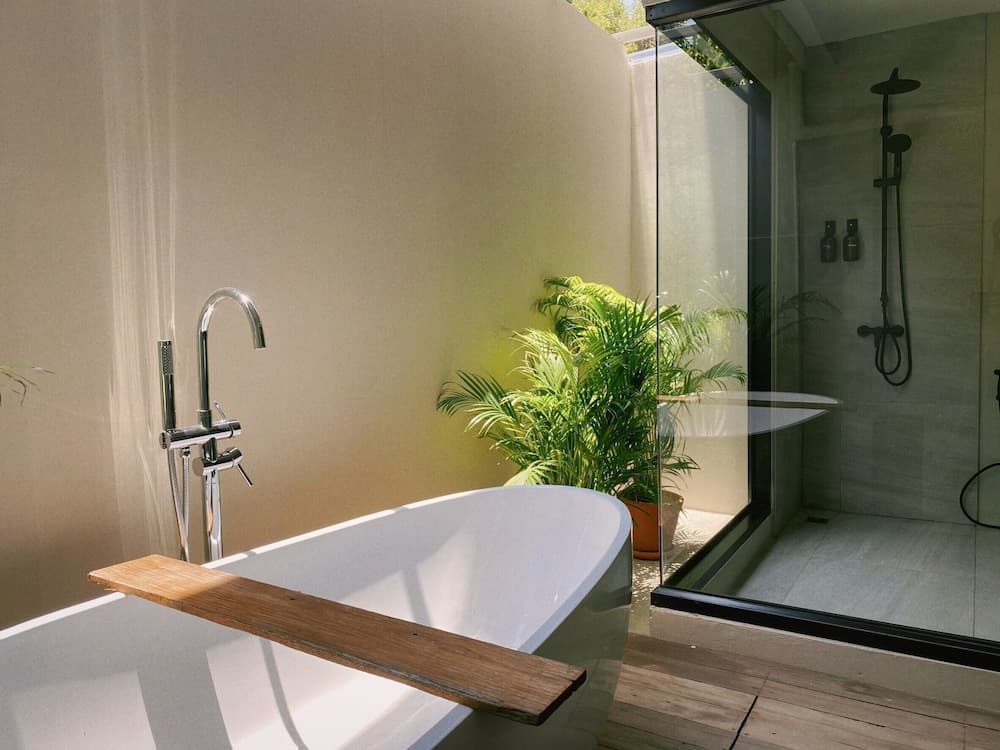 A serene outdoor bathroom features a modern bathtub with a wooden shelf, lush greenery, and a glass-enclosed shower, ideal for a tranquil spa retreat experience.