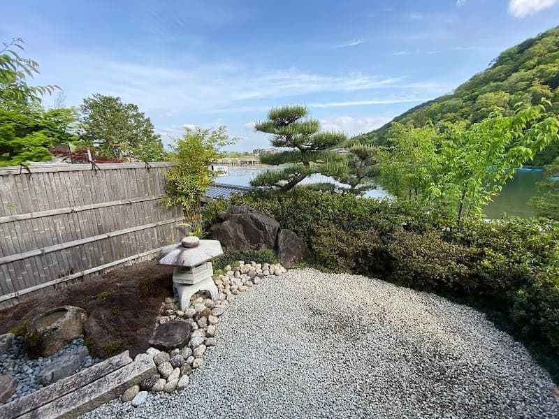 A serene Japanese garden featuring a stone lantern, gravel path, and lush greenery under a clear blue sky, ideal for tranquil meditation and cultural exploration in Kyoto's Ukyo Ward.