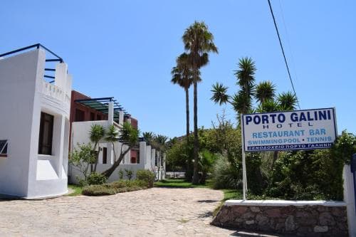 A sunny day at a tropical resort features white buildings, lush palm trees, and a welcoming sign for Porto Galini Hotel, ideal for travelers seeking relaxation and amenities like a restaurant, bar, swimming pool, and tennis courts.