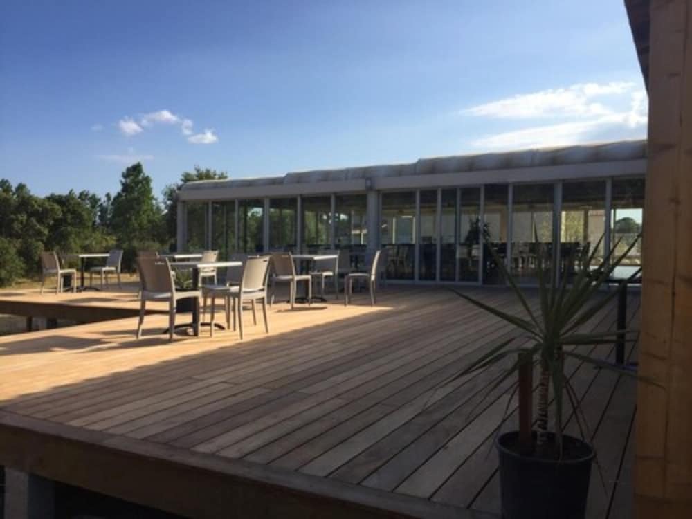 A serene outdoor dining terrace with wooden flooring, modern chairs, and tables under a clear blue sky, ideal for travelers seeking a relaxing meal in a tranquil setting near a glass-enclosed building.