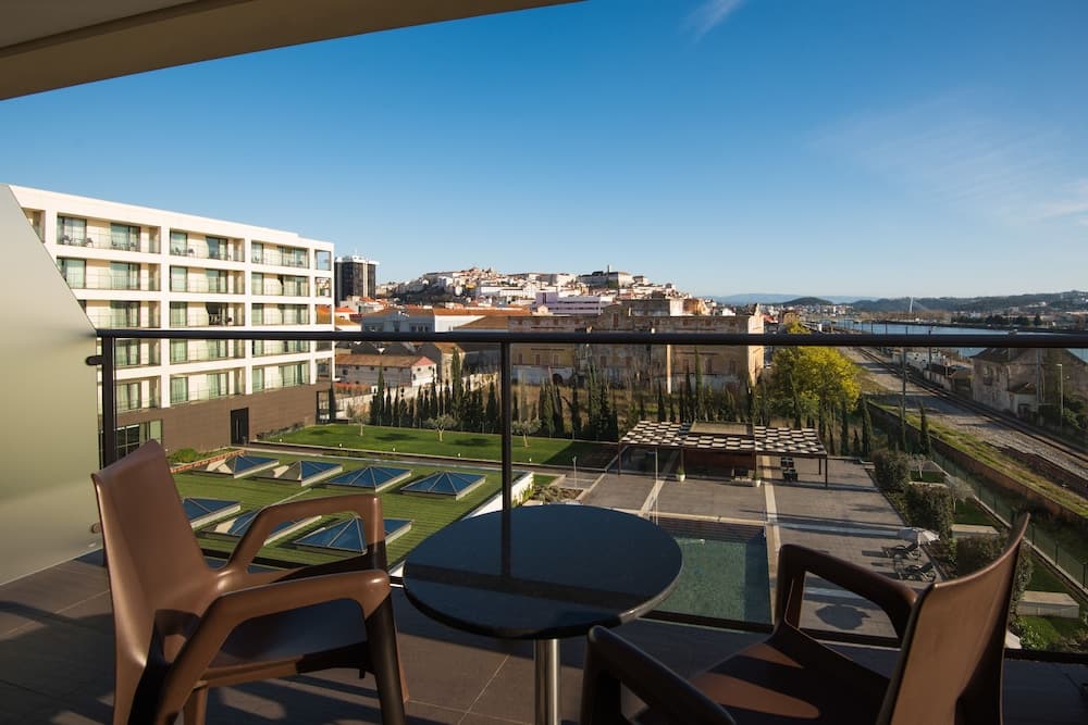 A serene balcony view showcases modern chairs and a table overlooking a lush green courtyard, contemporary buildings, and a distant cityscape under a clear blue sky, ideal for travelers seeking relaxation and urban exploration.