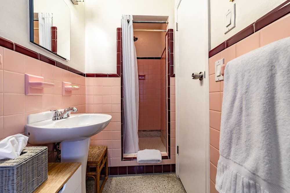 A cozy vintage bathroom with pink tiles, a white pedestal sink, and a shower area exudes a nostalgic charm, perfect for travelers seeking a retro-inspired accommodation experience.