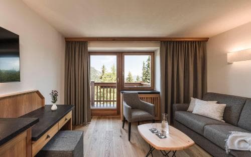 A cozy hotel suite with wooden floors, modern furnishings, and a balcony overlooking a scenic view, ideal for travelers seeking comfort and relaxation in a mountain retreat.