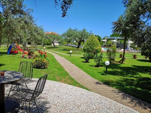A serene resort garden features lush greenery, colorful playground equipment, and a paved pathway leading to a pool area under clear blue skies, ideal for family vacations seeking relaxation and recreation.