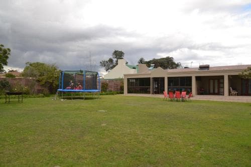 A serene outdoor space features a trampoline on a lush lawn beside a modern building with red chairs, ideal for family-friendly retreats under a cloudy sky.