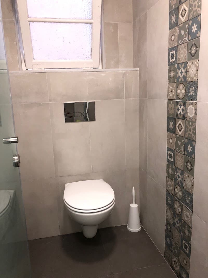A modern bathroom at the Vistas de Lisboa Hostel in Lisbon features sleek tiled walls, a wall-mounted toilet, decorative patterned tiles, and a frosted window, offering travelers a clean and stylish accommodation option.