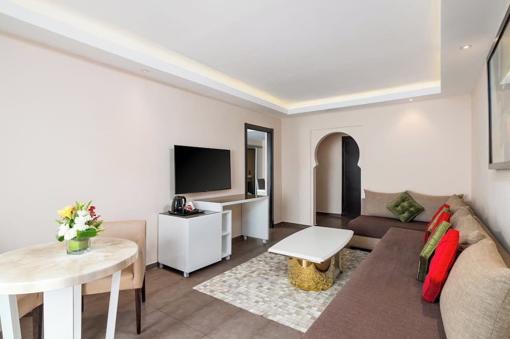 A cozy modern living room with a sleek sofa adorned with red and green cushions, a stylish coffee table, a flat-screen TV, and a dining area featuring a vase of fresh flowers, ideal for travelers seeking comfort in a boutique hotel suite.