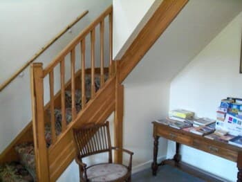 A cozy nook under a wooden staircase features a vintage chair and a table laden with books, creating an inviting spot for quiet reading or waiting in a quaint, homey setting.