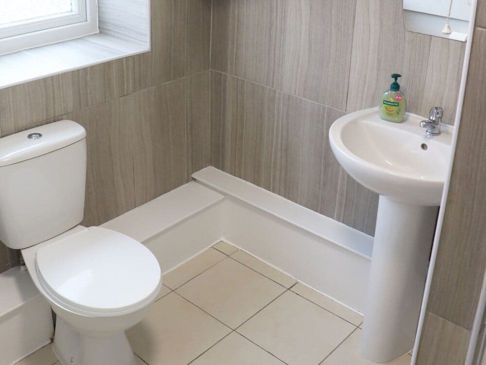A cozy bathroom with wood-patterned walls, a white toilet, pedestal sink, and tiled floor offers a clean and functional space ideal for travelers seeking comfort in accommodations.