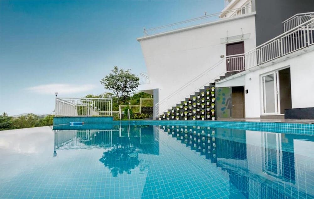 A serene resort pool area features a modern building with a staircase adorned with potted plants, surrounded by lush greenery, ideal for travelers seeking relaxation and luxury accommodations.