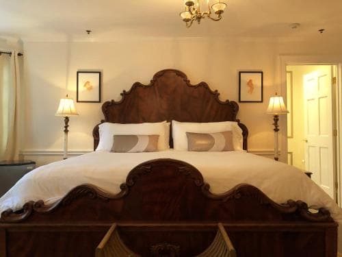 A cozy hotel room features an ornate wooden bed with plush white linens, framed by warm lighting from bedside lamps, creating an inviting atmosphere ideal for travelers seeking comfort and relaxation.
