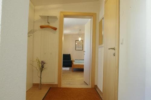 A cozy apartment hallway leads to a warmly lit bedroom, featuring simple decor, wooden accents, and a vase of flowers, ideal for travelers seeking comfortable and budget-friendly accommodation.