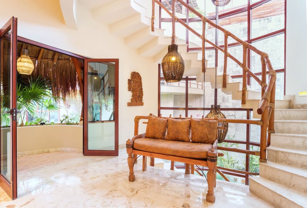 A luxurious tropical villa lobby features a wooden bench with plush cushions, a spiral staircase with bamboo railings, and large windows overlooking lush greenery, ideal for travelers seeking a serene and elegant retreat.