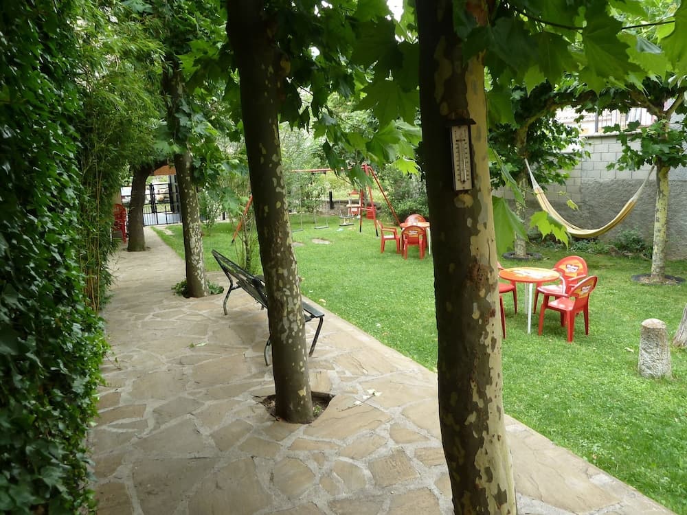 A serene garden pathway lined with lush trees leads to a vibrant play area featuring red chairs, a hammock, and swings, ideal for family-friendly outdoor relaxation and recreation.