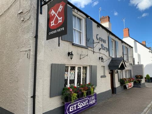 A charming village pub named "Cross Keys" with a welcoming facade, vibrant flower boxes, and a BT Sport sign, offers a cozy atmosphere perfect for travelers seeking a traditional British drinking spot.