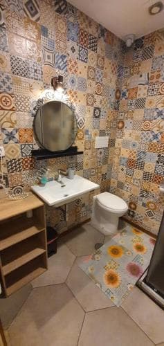 A cozy bathroom with vibrant patchwork tiles exudes charm, featuring a sleek sink, wooden shelving, and a floral rug, ideal for travelers seeking unique accommodation aesthetics.