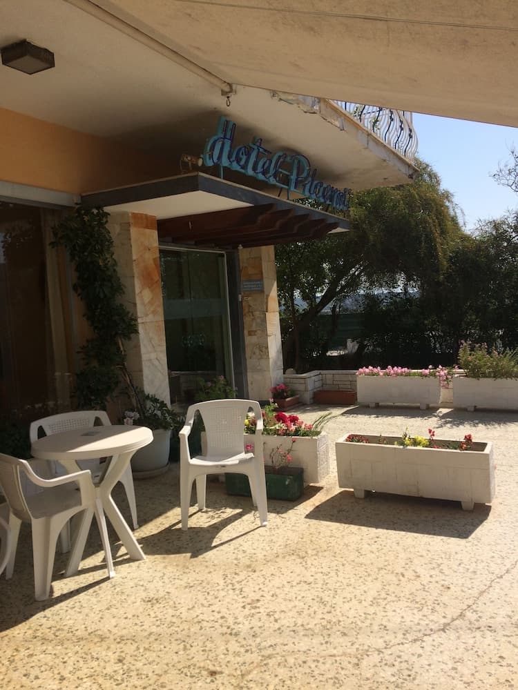 A serene hotel patio features white plastic furniture, vibrant flower planters, and a blue neon sign under a sunny sky, ideal for travelers seeking relaxation in a cozy outdoor setting.