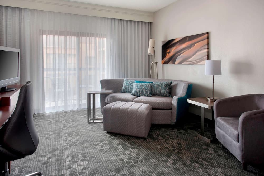 A cozy hotel suite features a comfortable living area with a sofa, armchair, desk, and large window draped in sheer curtains, creating a serene atmosphere ideal for relaxation or business travelers.