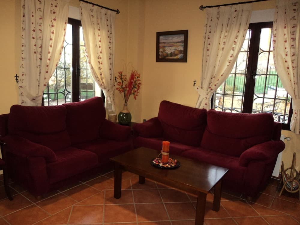 A cozy living room with terracotta flooring, plush red sofas, floral curtains, and a wooden coffee table creates an inviting atmosphere ideal for travelers seeking a comfortable retreat.
