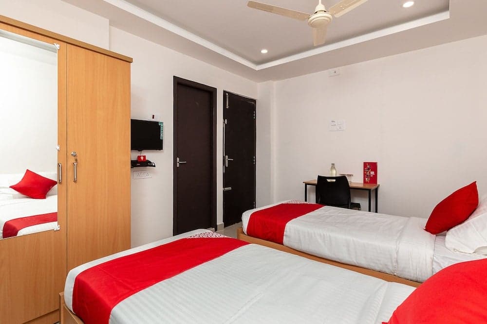 A cozy hotel room features two neatly made beds with red accents, a wooden wardrobe, a small desk area, and a mounted TV, ideal for travelers seeking comfort and functionality.