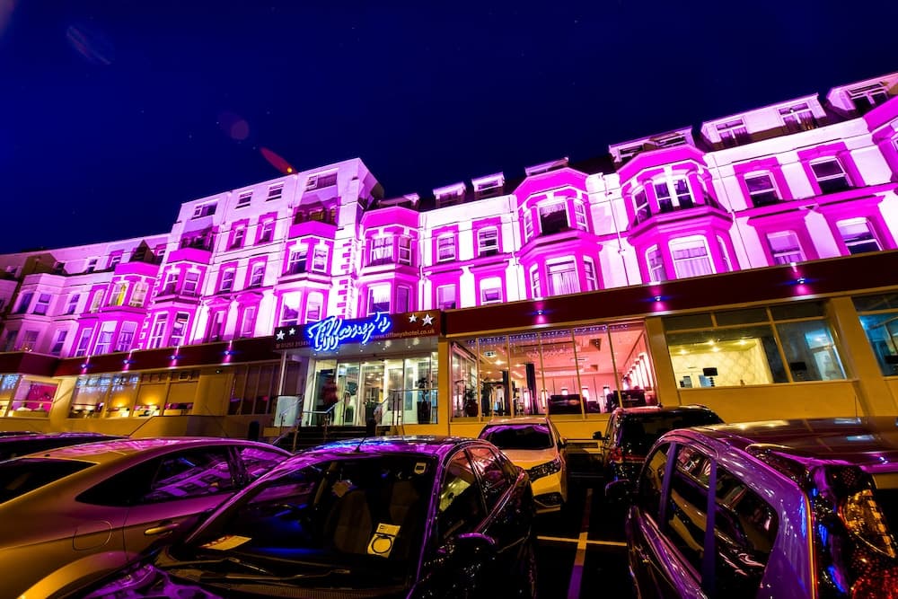A vibrant nighttime urban scene features a grand building illuminated in pink hues, with parked cars reflecting the glow, ideal for travelers seeking lively city nightlife experiences.