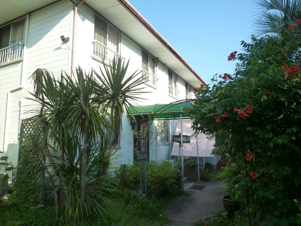 A serene residential garden with lush greenery, vibrant red flowers, and a palm tree frames the entrance to a quaint house, ideal for travelers seeking a peaceful retreat in a suburban setting.